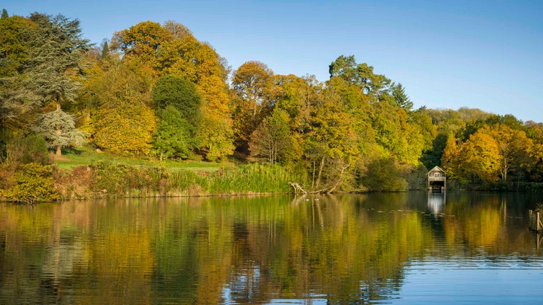 A view across a lake towards a small boathouse at Winkworth Arboretum, Surrey. Many trees stand on the bank of the lake and their leaves are beginning to turn yellow and orange.